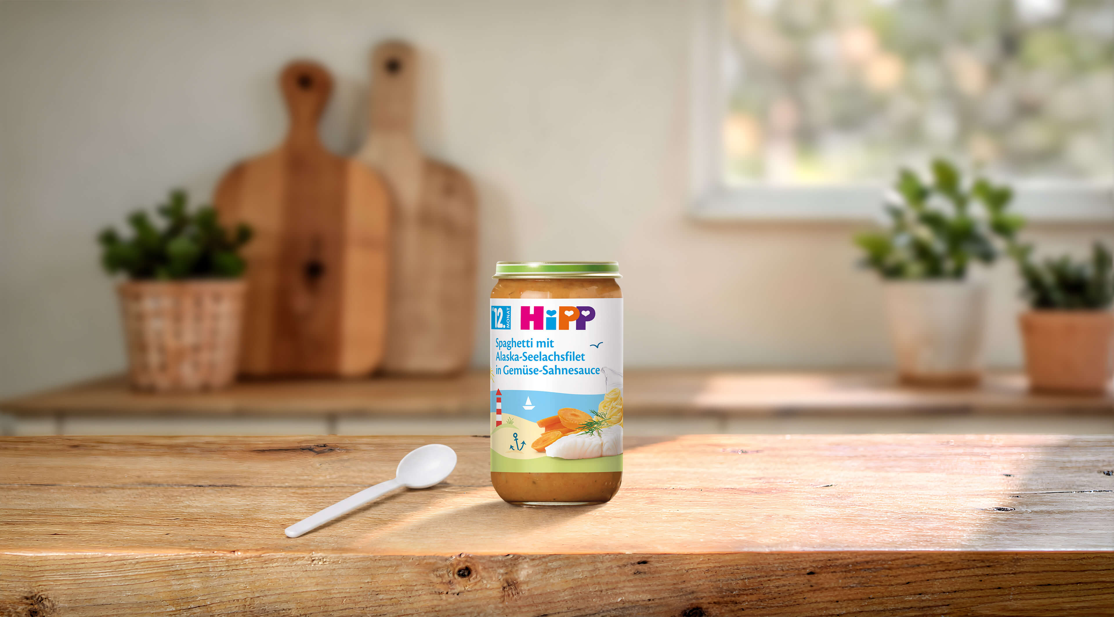 HiPP Bio Babyglas Spaghetti mit Alaska-Seelachsfilet in Gemüse-Sahnesauce ab 12 Monaten auf Holztisch mit Löffel in heller Küche