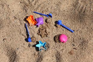 Mehrere blaue Milchpulverlöffel und bunte Sandförmchen liegen im Sandkasten und formen kleine Sandsterne und -muscheln.