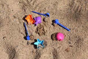 Mehrere blaue Milchpulverlöffel und bunte Sandförmchen liegen im Sandkasten und formen kleine Sandsterne und -muscheln.