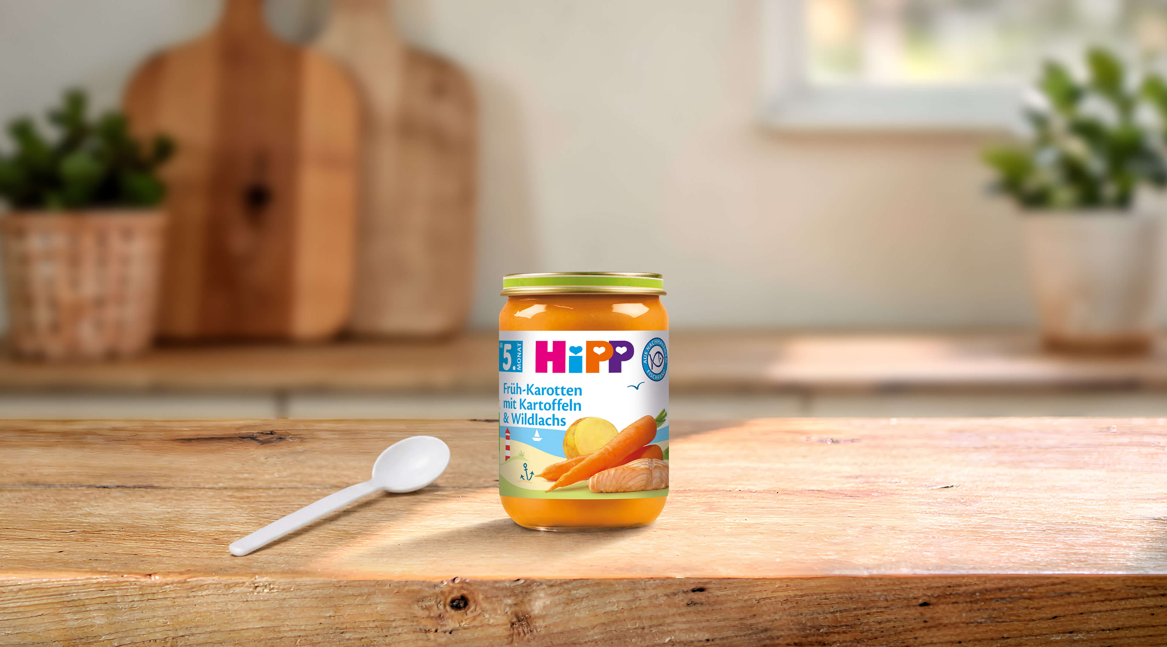 HiPP Früh-Karotten mit Kartoffeln & Wildlachs Bio-Babygläschen auf Holztisch mit Löffel in heller Küche