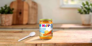 HiPP Früh-Karotten mit Kartoffeln & Wildlachs Bio-Babygläschen auf Holztisch mit Löffel in heller Küche
