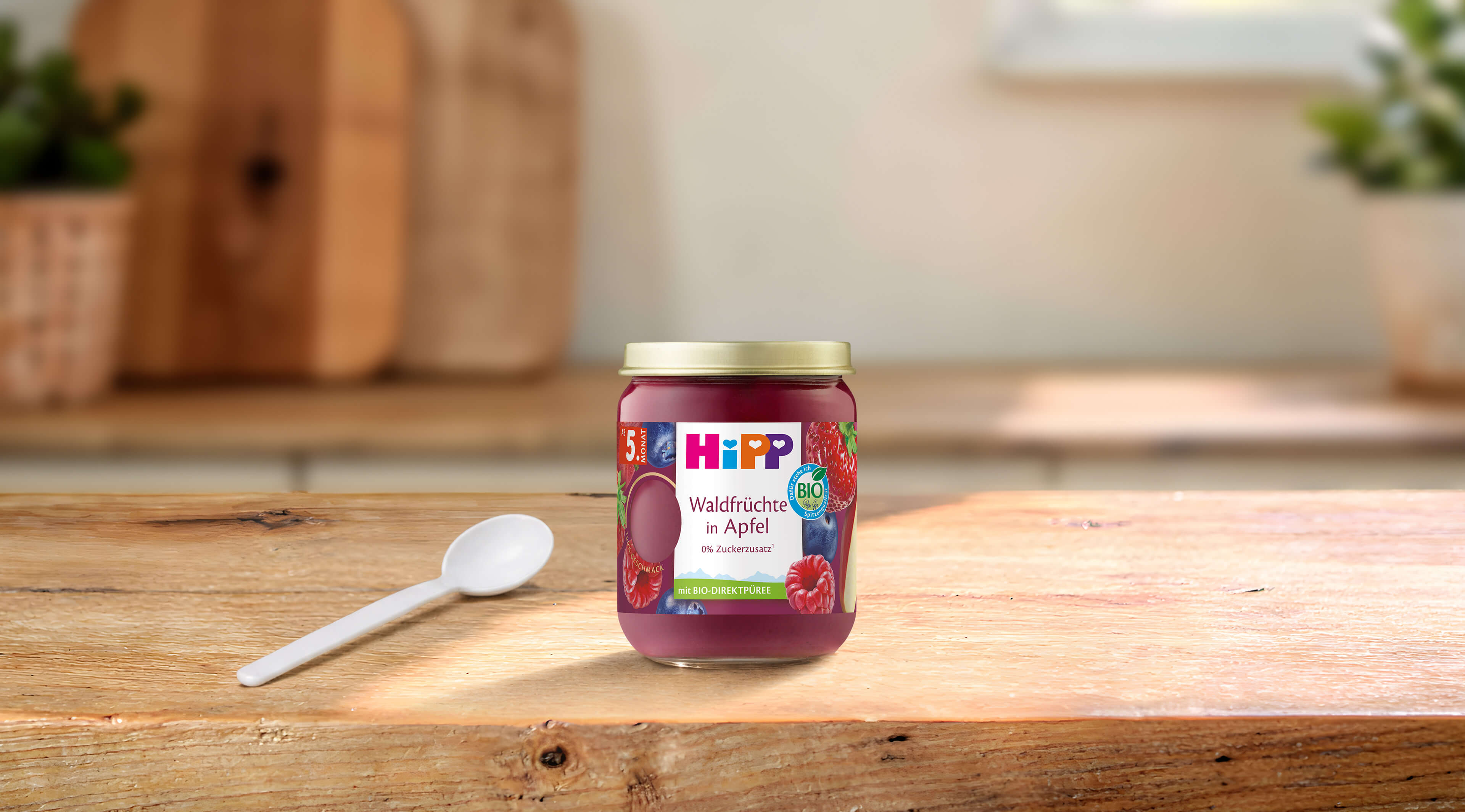 HiPP Bio-Gläschen „Waldfrüchte in Apfel“ ab 5. Monat auf Holztisch, daneben ein kleiner Löffel in heller Küchenumgebung