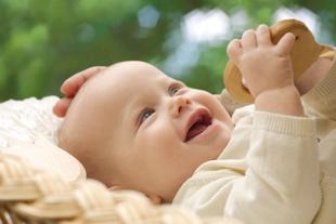 Ein fröhliches, weiß gekleidetes Baby spielt mit einem Holzspielzeug, während es in einem geflochtenen Korb gewiegt wird.