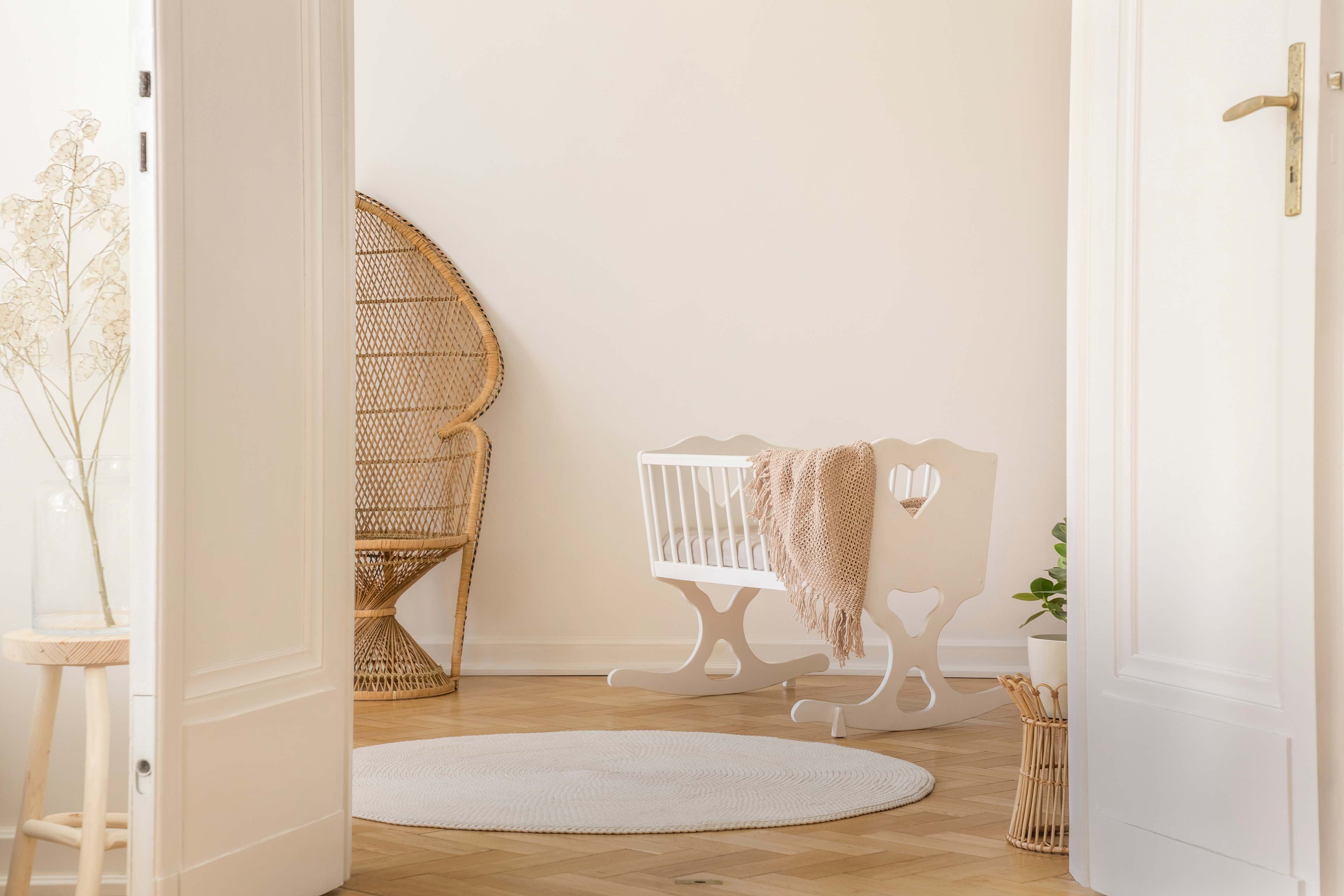 Ein ruhiges Zimmer mit einem Korbsessel, einem weißen Kinderbett, einem runden Teppich und einer hohen Vase mit getrockneten Pflanzen.