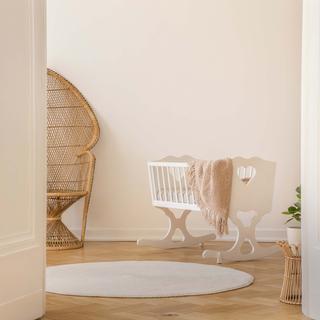 Ein ruhiges Zimmer mit einem Korbsessel, einem weißen Kinderbett, einem runden Teppich und einer hohen Vase mit getrockneten Pflanzen.