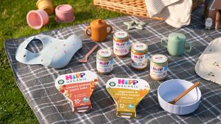HiPP Organic baby food on a picnic blanket with a spoon, bowl, bib, sippy cup and wicker basket
