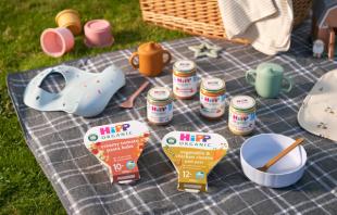 HiPP Organic baby food on a picnic blanket with a spoon, bowl, bib, sippy cup and wicker basket