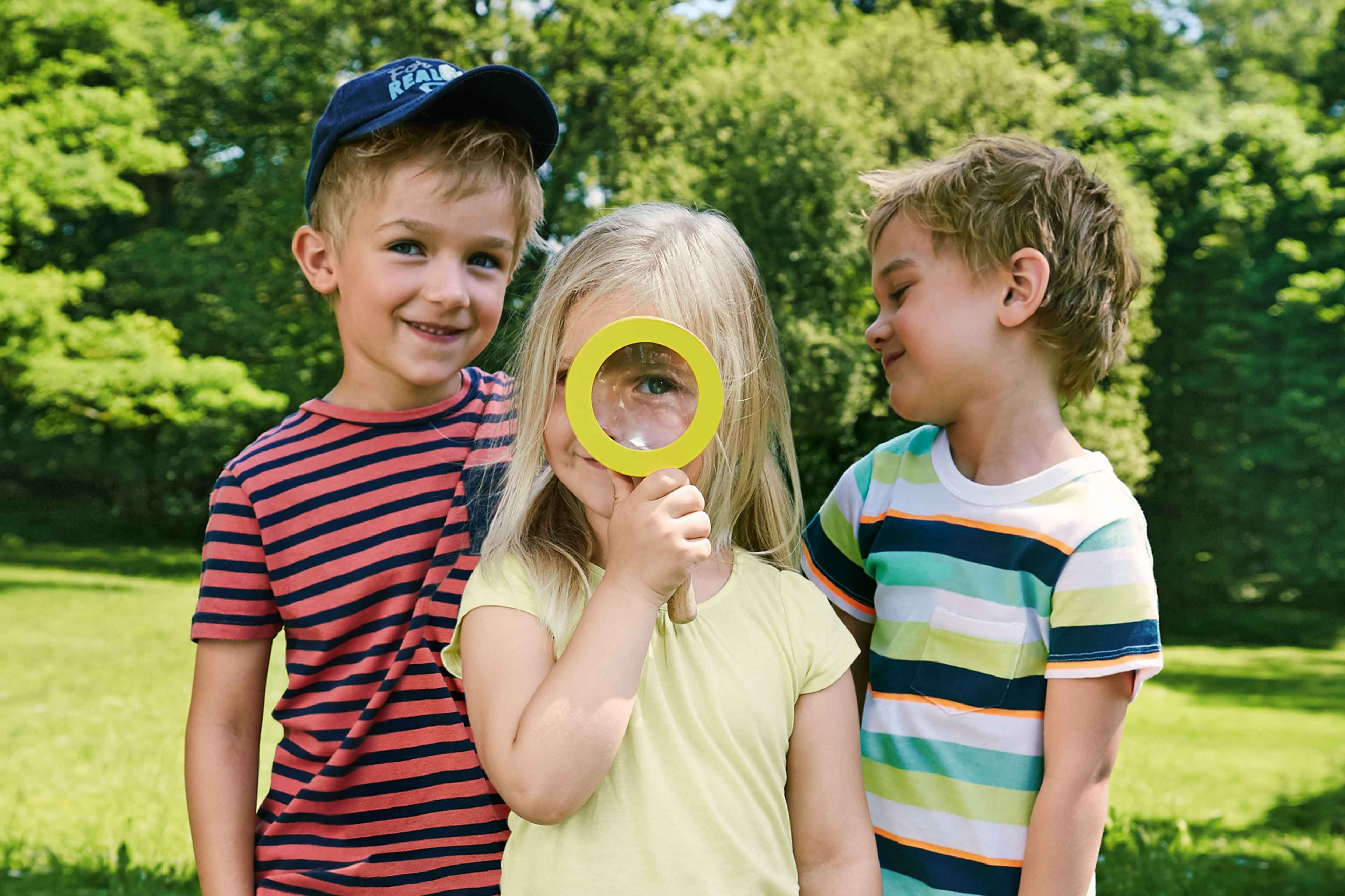 Drei Kinder in einem Park, von denen eines ein gelbes Vergrößerungsglas über sein Auge hält.