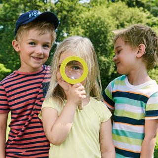Drei Kinder in einem Park, von denen eines ein gelbes Vergrößerungsglas über sein Auge hält.