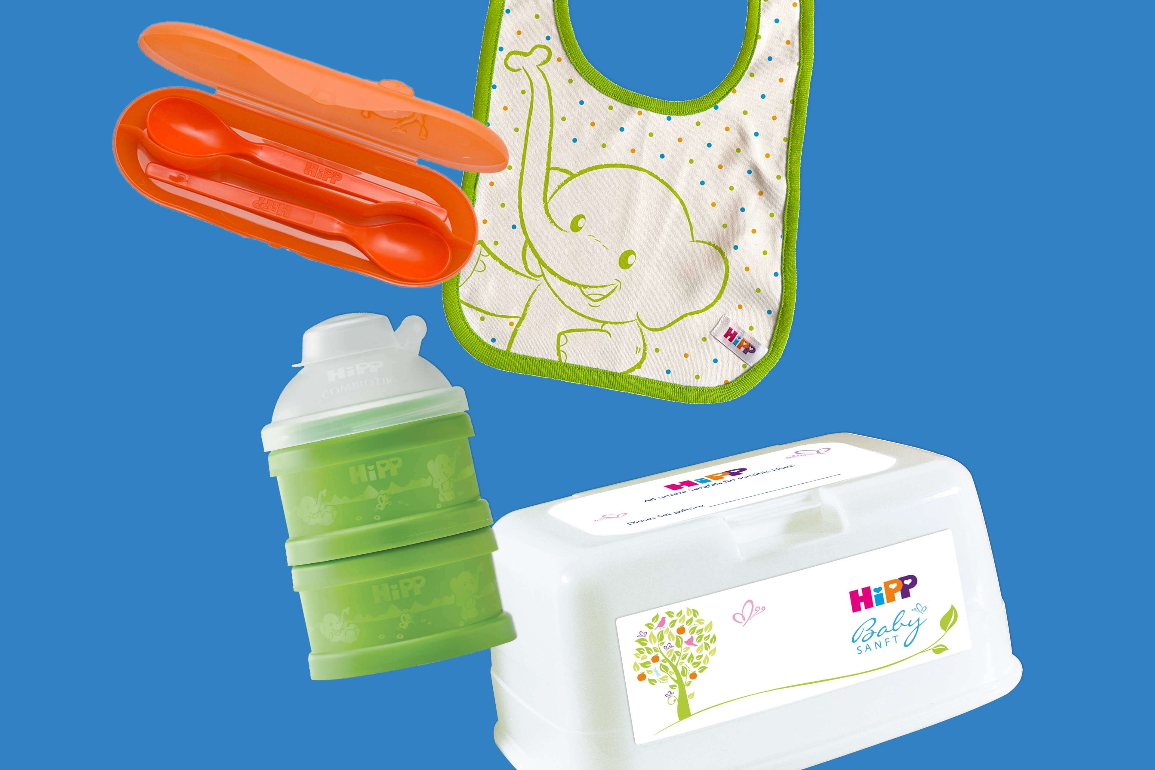 Grüner Babynahrung-Portionierer, orangefarbene Löffel in passender Box, weißer Feuchttücherbehälter mit HiPP-Logo und grüner Baumillustration sowie weiß-grüner Lätzchen mit Elefantenmotiv vor blauem Hintergrund