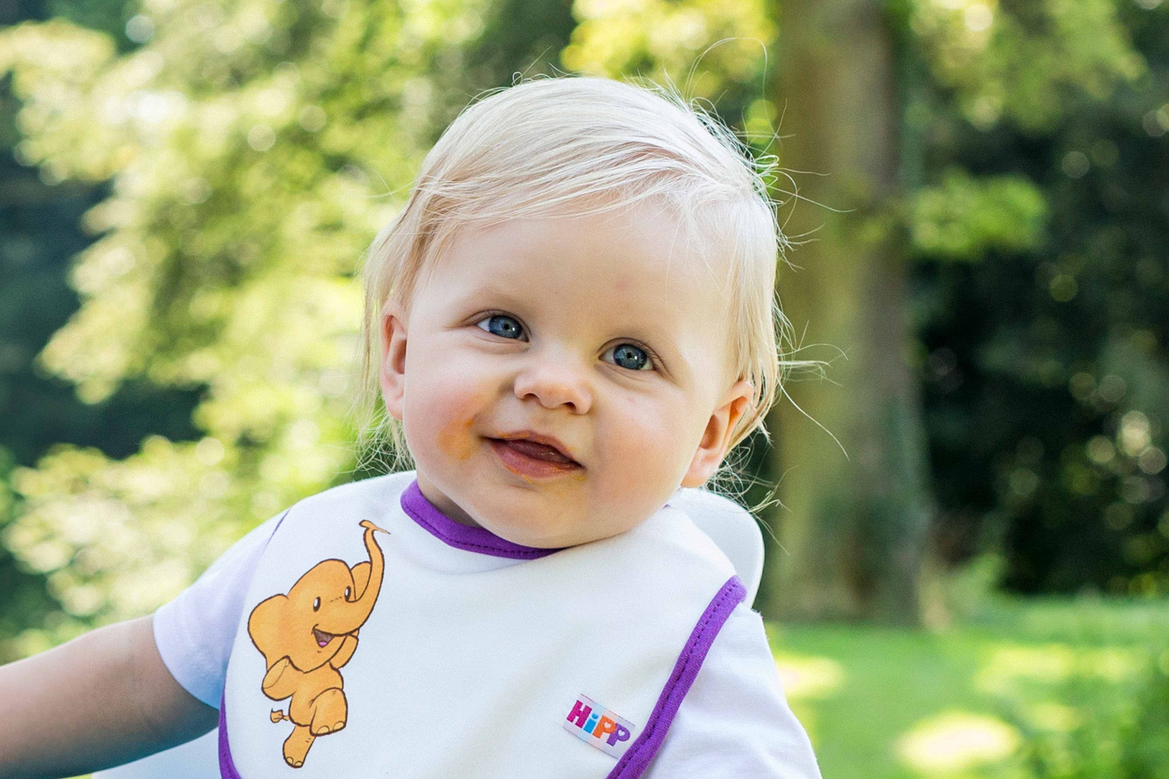 Ein Baby mit blondem Haar, blauen Augen und einem Lätzchen mit Elefantenmotiv vor einem grünen Hintergrund.