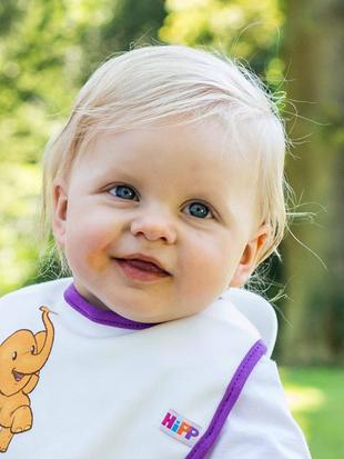 Ein Baby mit blondem Haar, blauen Augen und einem Lätzchen mit Elefantenmotiv vor einem grünen Hintergrund.