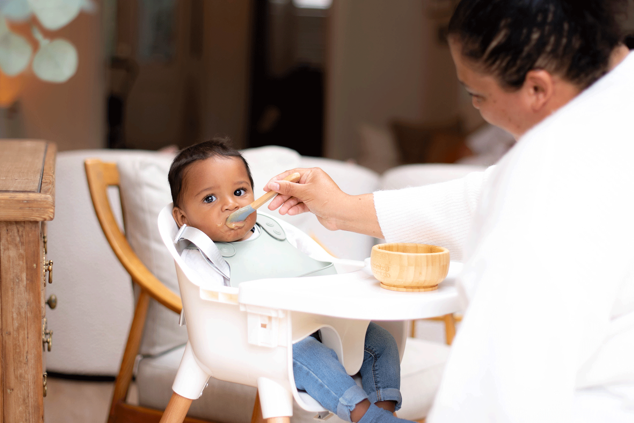 A baby wearing a sage green bib and denim jeans sits in a white high chair while an adult feeds them pureed food with a wooden spoon. On the high chair tray sits a small wooden bowl, and a softly blurred living room is visible in the background.