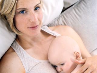 Frau mit kurzen blonden Haaren liegt auf einem Kissen und hält ein Baby im Arm