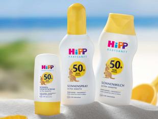 Drei Sonnenschutzprodukte der Marke "HiPP" mit unterschiedlichem Lichtschutzfaktor, platziert an einem Sandstrand mit einem klaren blauen Himmel im Hintergrund.