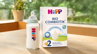 HiPP 2 BIO COMBIOTIK Folgemilch-Packung mit gefüllter Babyflasche auf Holztisch, daneben blauer Kreis mit „Jetzt kostenlos testen!“