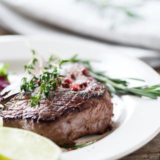 Ein gegrilltes Stück Fleisch, garniert mit Kräutern und einer Limettenscheibe, begleitet von einer Beilage aus grünem Gemüse.