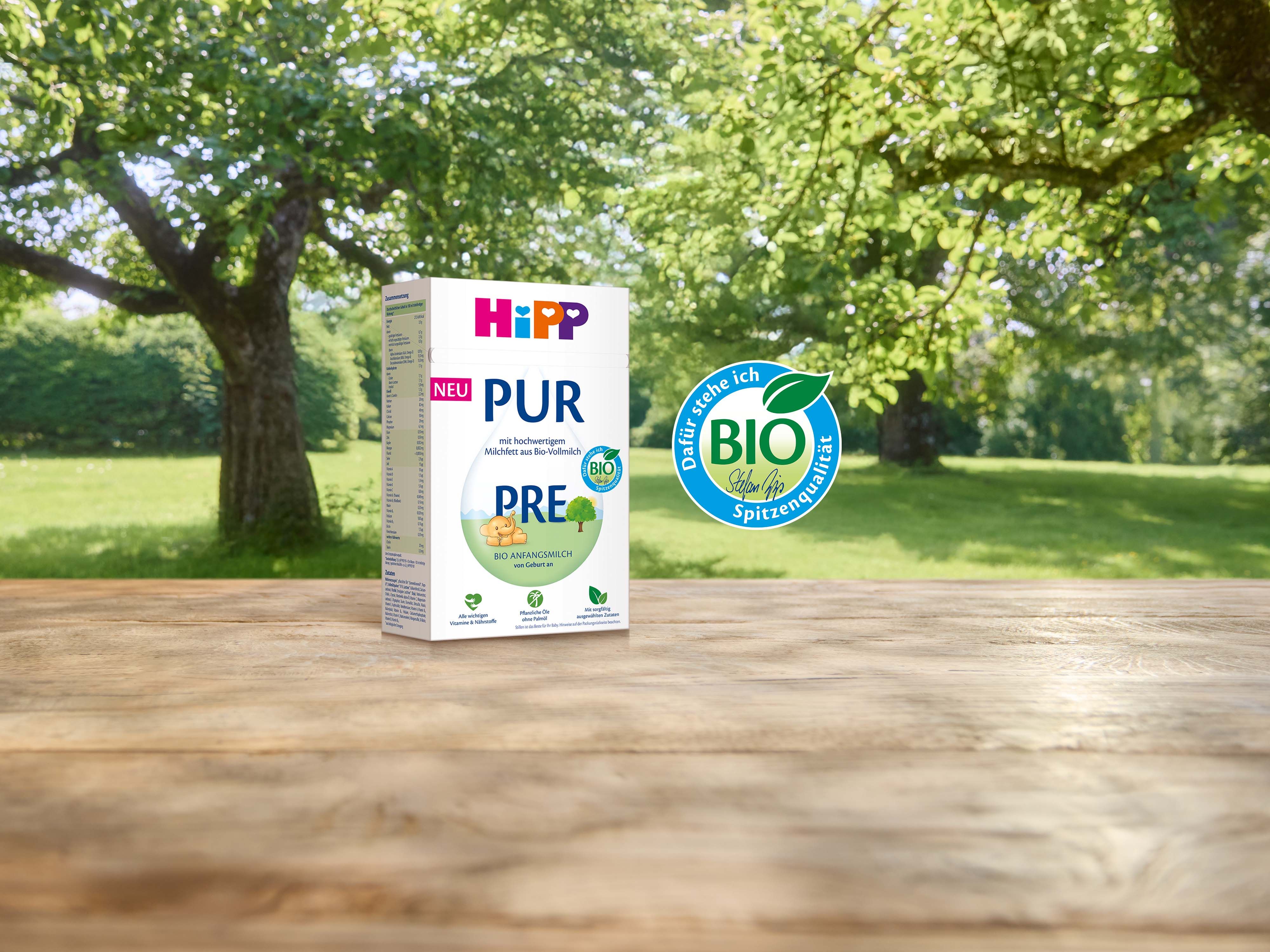 HiPP PRE PUR Bio-Anfangsmilch Packung auf Holztisch vor grüner Wiese mit großem Baum, daneben das HiPP Bio-Siegel..