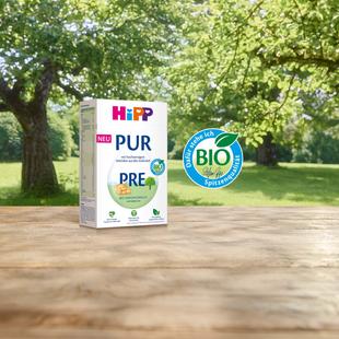 HiPP PRE PUR Bio-Anfangsmilch Packung auf Holztisch vor grüner Wiese mit großem Baum, daneben das HiPP Bio-Siegel..