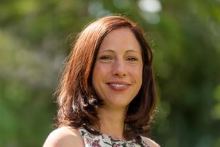 Eine Frau mit braunem Haar, die ein geblümtes Oberteil trägt, lächelt und schaut direkt in die Kamera.