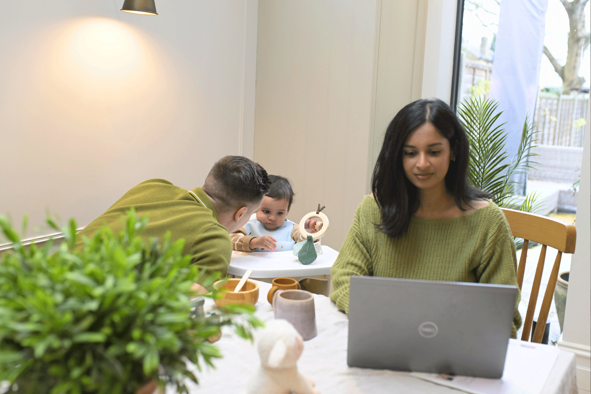 A woman sits at a dining table working on a grey laptop, while a man next to her leans over to interact with a baby sitting in a high chair. The baby, wearing a bib, plays with a wooden ring toy. On the table, in front of the laptop, are wooden bowls, mugs, and a small plush sheep toy.