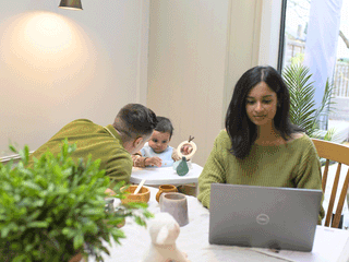 A woman sits at a dining table working on a grey laptop, while a man next to her leans over to interact with a baby sitting in a high chair. The baby, wearing a bib, plays with a wooden ring toy. On the table, in front of the laptop, are wooden bowls, mugs, and a small plush sheep toy.