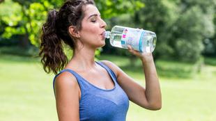 Eine Frau in einem blauen Tank-Top trinkt Wasser aus einer Flasche mit der Aufschrift "Eau de France" in einer Außenumgebung mit Grün im Hintergrund.