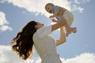 Lachende Mutter hebt ihr fröhliches Baby im Freien in die Luft, beide genießen gemeinsam die frische Luft und Nähe