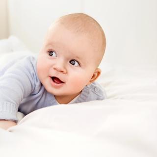 Lächelndes Baby in hellblauem Pulli liegt auf dem Bauch im Bett – Symbol für die Suche nach einem schönen Jungennamen