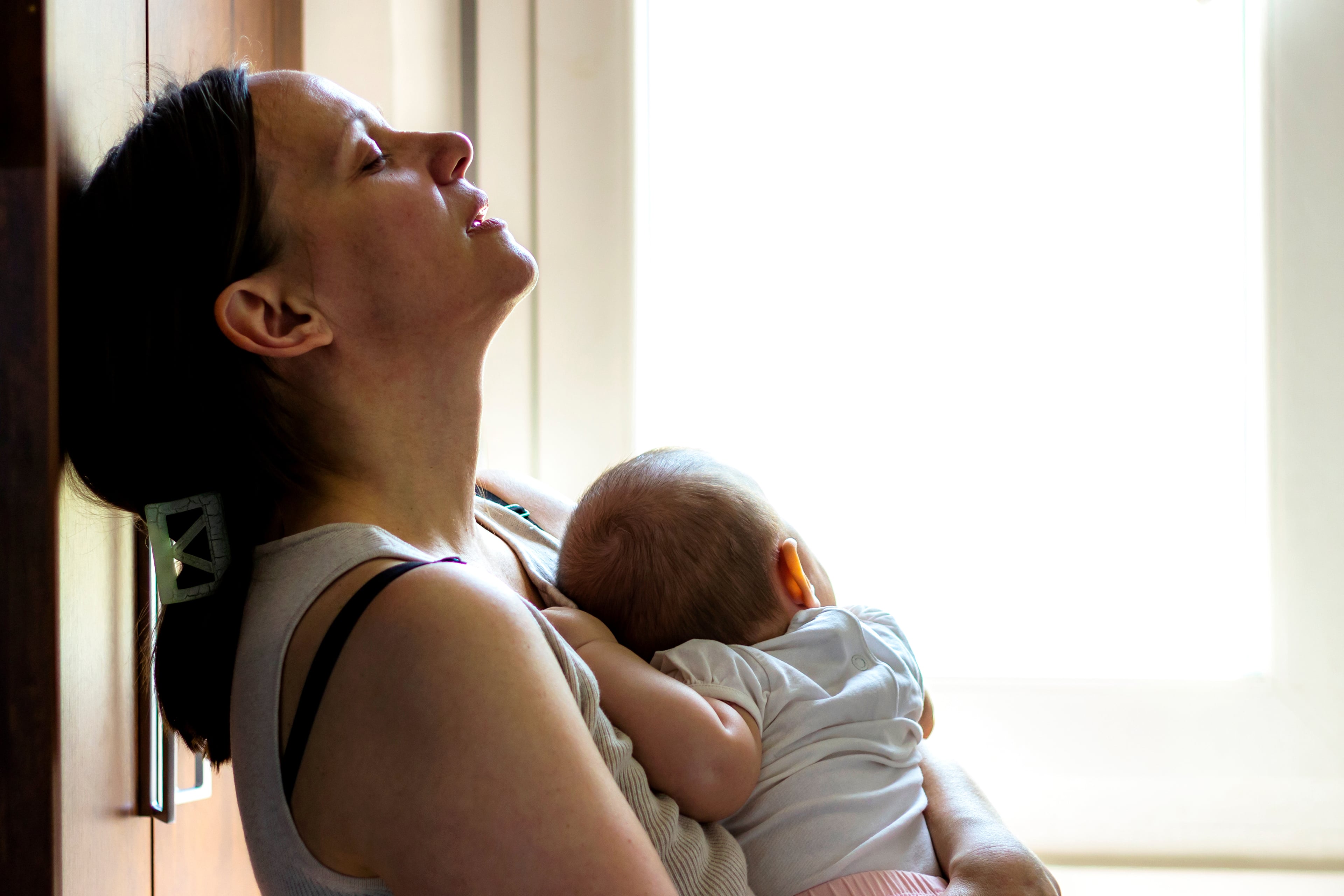 Eine Frau mit geschlossenen Augen, die nach oben blickt und ein Baby an ihrer Brust hält, wobei ein Fenster die Szene beleuchtet.