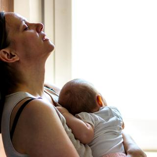 Eine Frau mit geschlossenen Augen, die nach oben blickt und ein Baby an ihrer Brust hält, wobei ein Fenster die Szene beleuchtet.