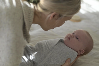 A woman gently cradles a swaddled baby in a soft grey onesie, gazing closely into the baby’s eyes during a quiet bonding moment. Natural light softly filters in, adding warmth to the serene setting