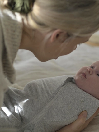 A woman gently cradles a swaddled baby in a soft grey onesie, gazing closely into the baby’s eyes during a quiet bonding moment. Natural light softly filters in, adding warmth to the serene setting.