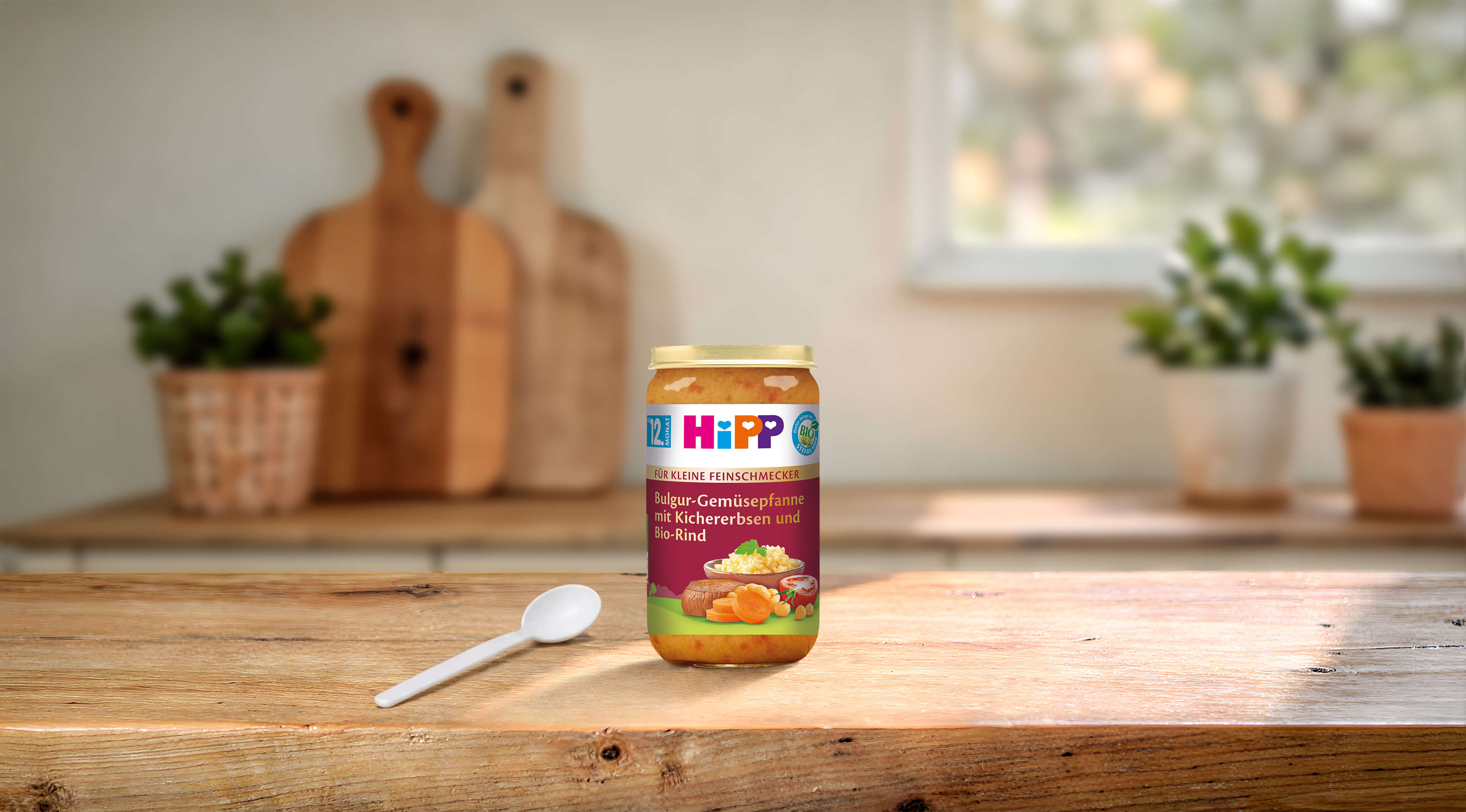 HiPP Glas Bulgur-Gemüsepfanne mit Kichererbsen und Bio-Rind auf Holztisch in heller, gemütlicher Küche mit Löffel daneben
