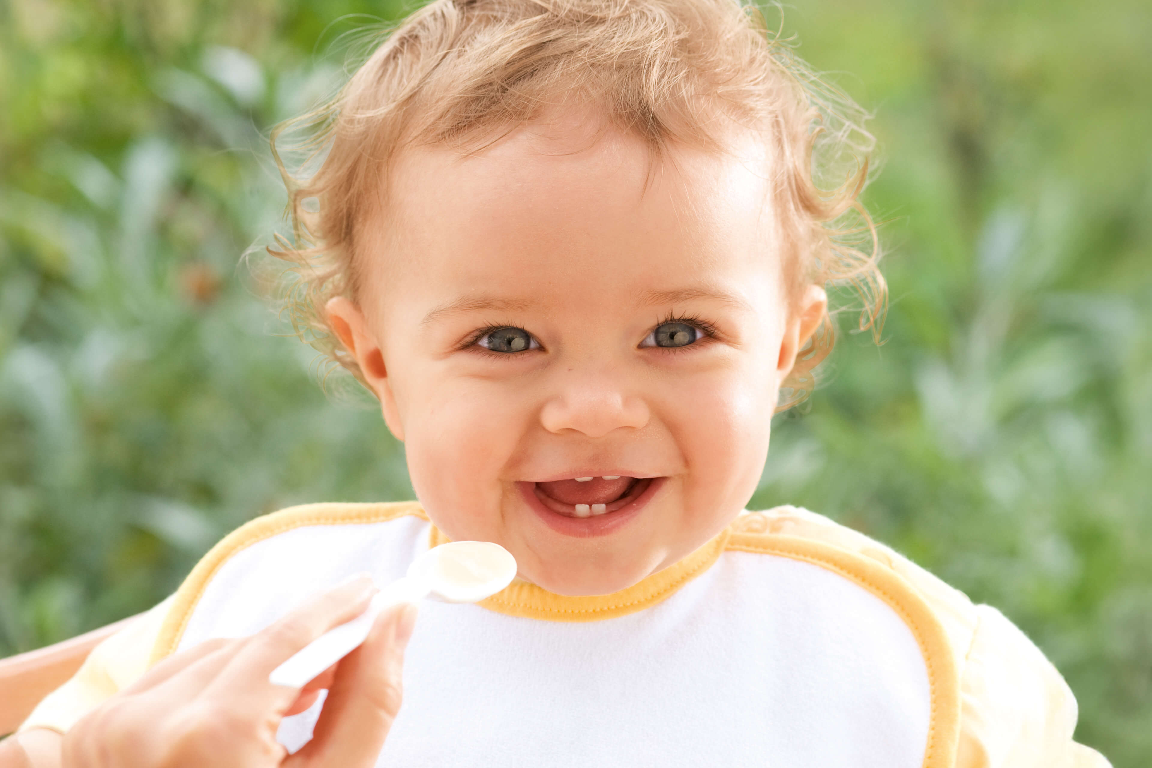Ein fröhliches Baby mit lockigem Haar, das ein Lätzchen trägt, lächelt und direkt in die Kamera schaut, während es mit einem Löffel gefüttert wird.