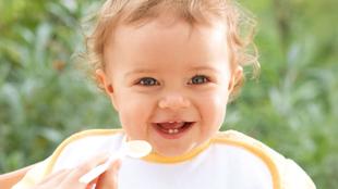 Ein fröhliches Baby mit lockigem Haar, das ein Lätzchen trägt, lächelt und direkt in die Kamera schaut, während es mit einem Löffel gefüttert wird.