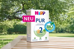 Packung HiPP PUR Bio-Folgemilch mit Aufschrift 'NEU' und Bio-Siegel auf Holztisch vor grünem Baumhintergrund