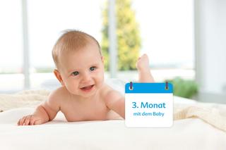 Ein vermutlich 3 Monate altes Baby liegt mit einem fröhlichen Gesichtsausdruck auf einem Bett, daneben ein Kalender mit der Aufschrift "3. Monat mit dem Baby".