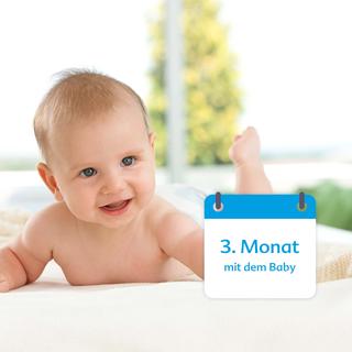 Ein vermutlich 3 Monate altes Baby liegt mit einem fröhlichen Gesichtsausdruck auf einem Bett, daneben ein Kalender mit der Aufschrift "3. Monat mit dem Baby".