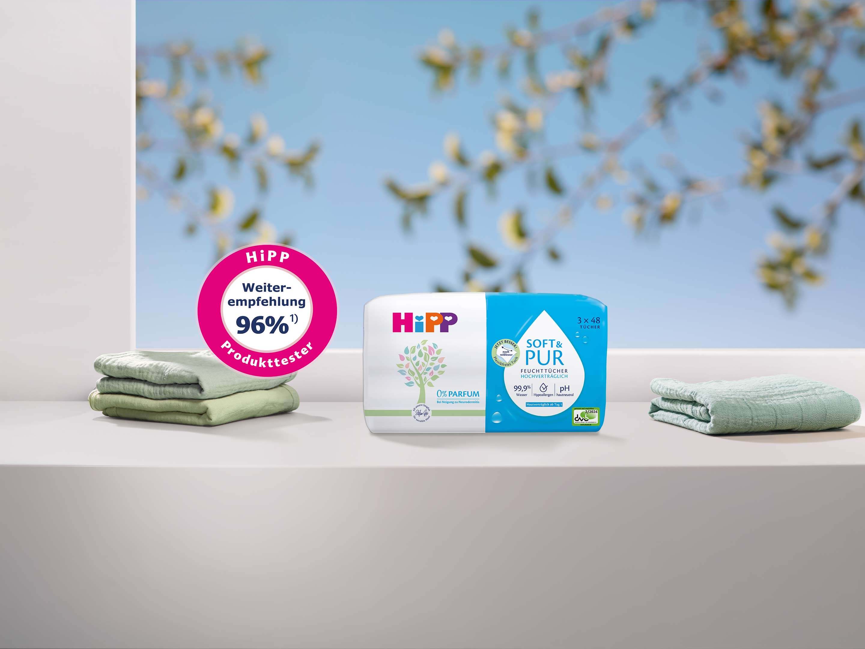 Zwei Babyproduktverpackungen von HiPP auf einer weißen Oberfläche mit zwei gefalteten Handtüchern links und rechts