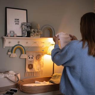 Woman standing in a softly lit nursery, holding up a tiny white baby onesie while preparing a changing station decorated with rainbows, stuffed animals, and ultrasound photos.