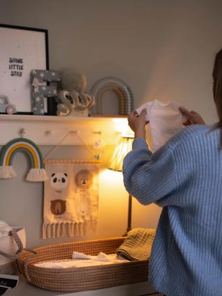 Woman standing in a softly lit nursery, holding up a tiny white baby onesie while preparing a changing station decorated with rainbows, stuffed animals, and ultrasound photos.