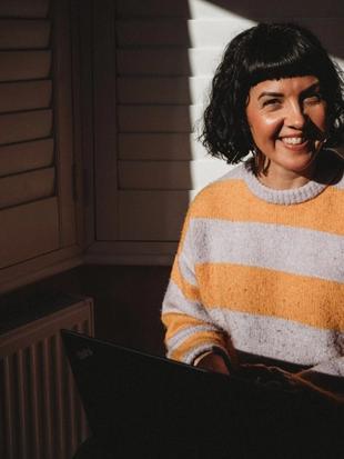 Photo of Lucy Wood, Paediatric Dietitian for HiPP Organic, smiling at the camera whilst working on a laptop with sunshine coming through a window on her face