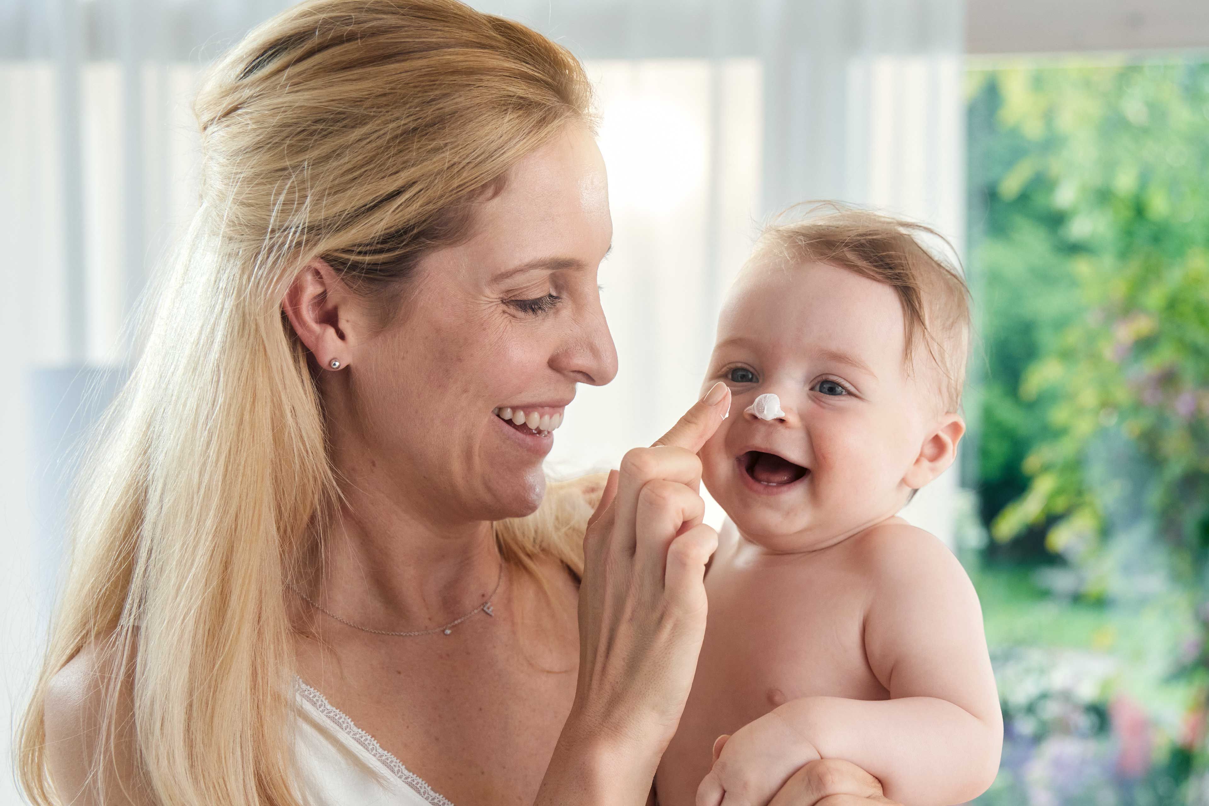 rau mit langem blondem Haar hält nacktes Baby vor hellem Fenster mit grünem Garten im Hintergrund
