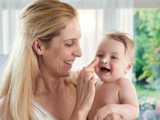 rau mit langem blondem Haar hält nacktes Baby vor hellem Fenster mit grünem Garten im Hintergrund