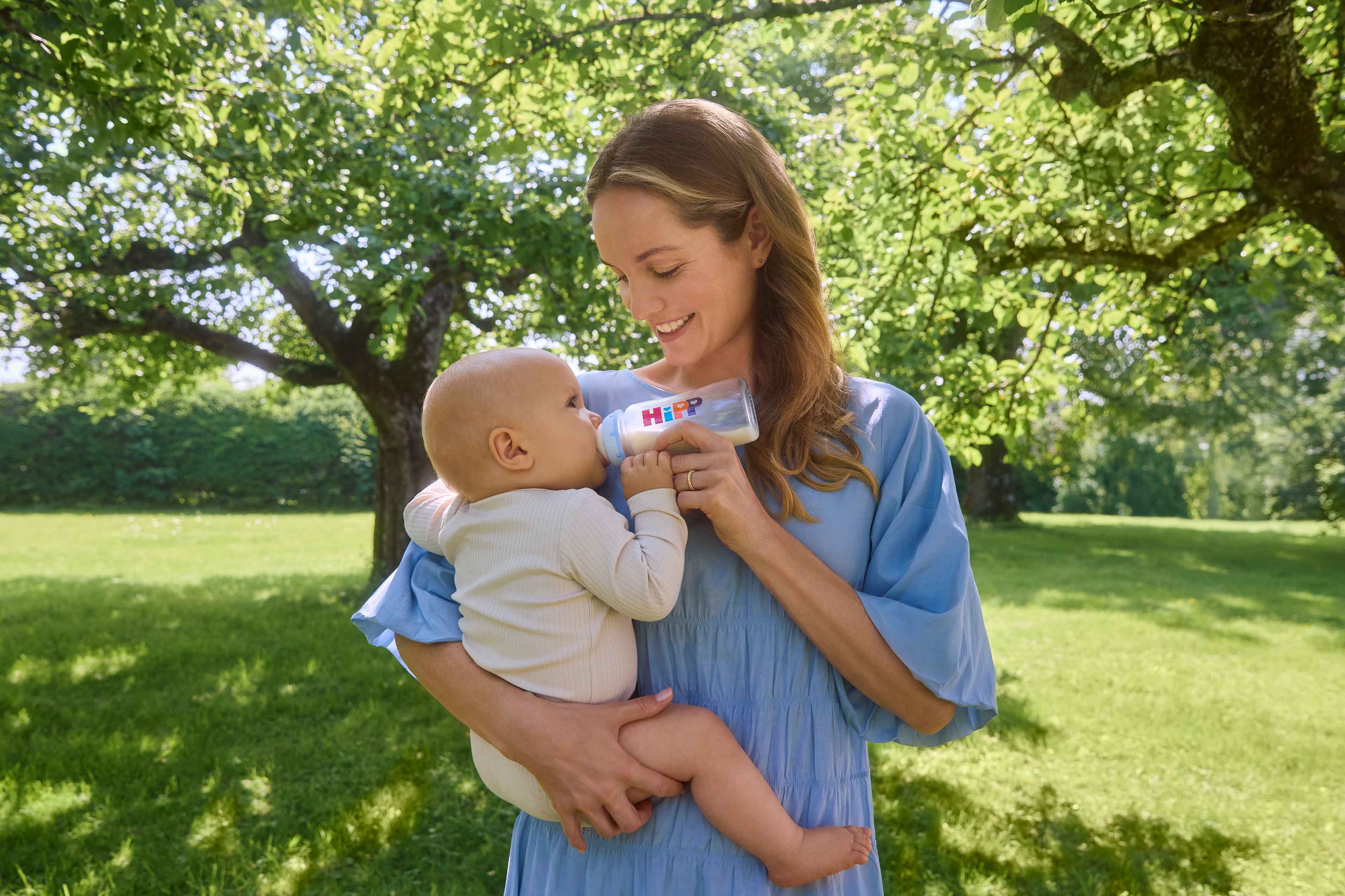 Lächelnde Mutter füttert ihr Baby im Arm mit einer HiPP Milchflasche in einem sonnigen, grünen Garten.