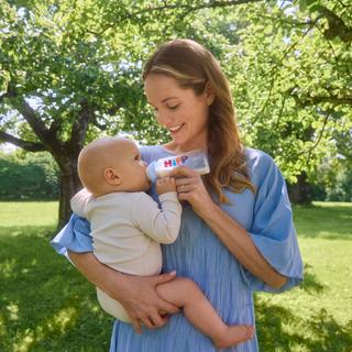 Lächelnde Mutter füttert ihr Baby im Arm mit einer HiPP Milchflasche in einem sonnigen, grünen Garten.