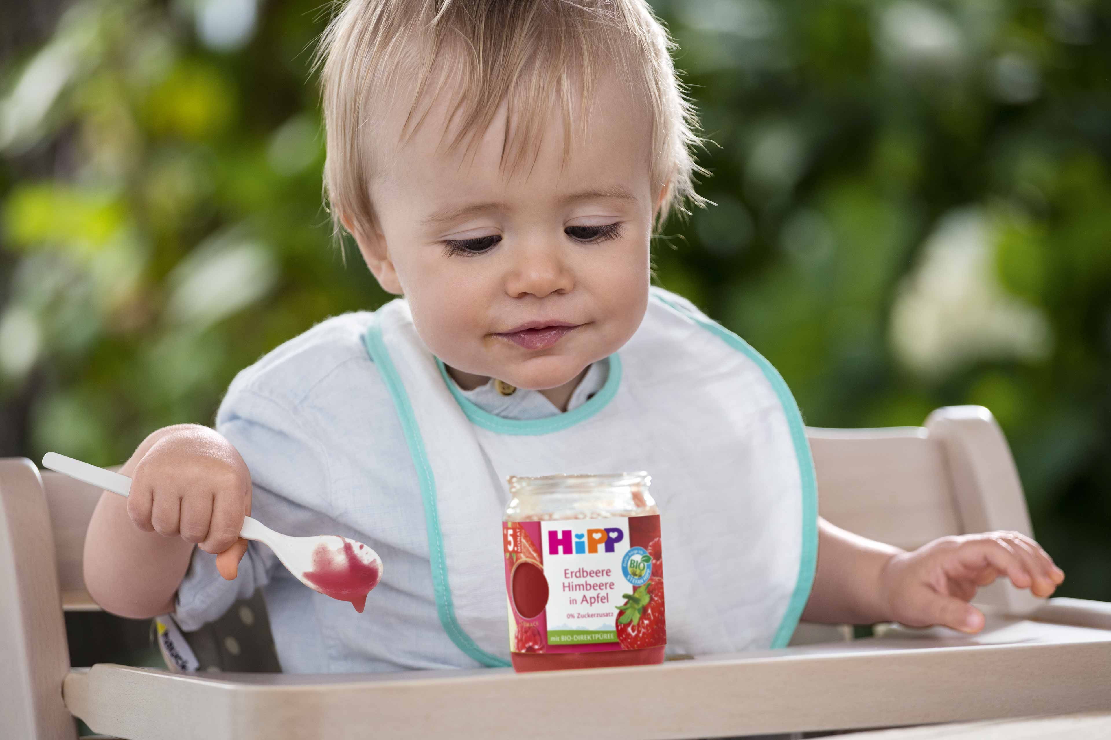 Ein kleines Kind, das in einem Hochstuhl sitzt, einen Löffel in der Hand hält und auf ein Glas mit Babynahrung mit der Aufschrift "HiPP" schaut.