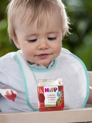 Ein kleines Kind, das in einem Hochstuhl sitzt, einen Löffel in der Hand hält und auf ein Glas mit Babynahrung mit der Aufschrift "HiPP" schaut.