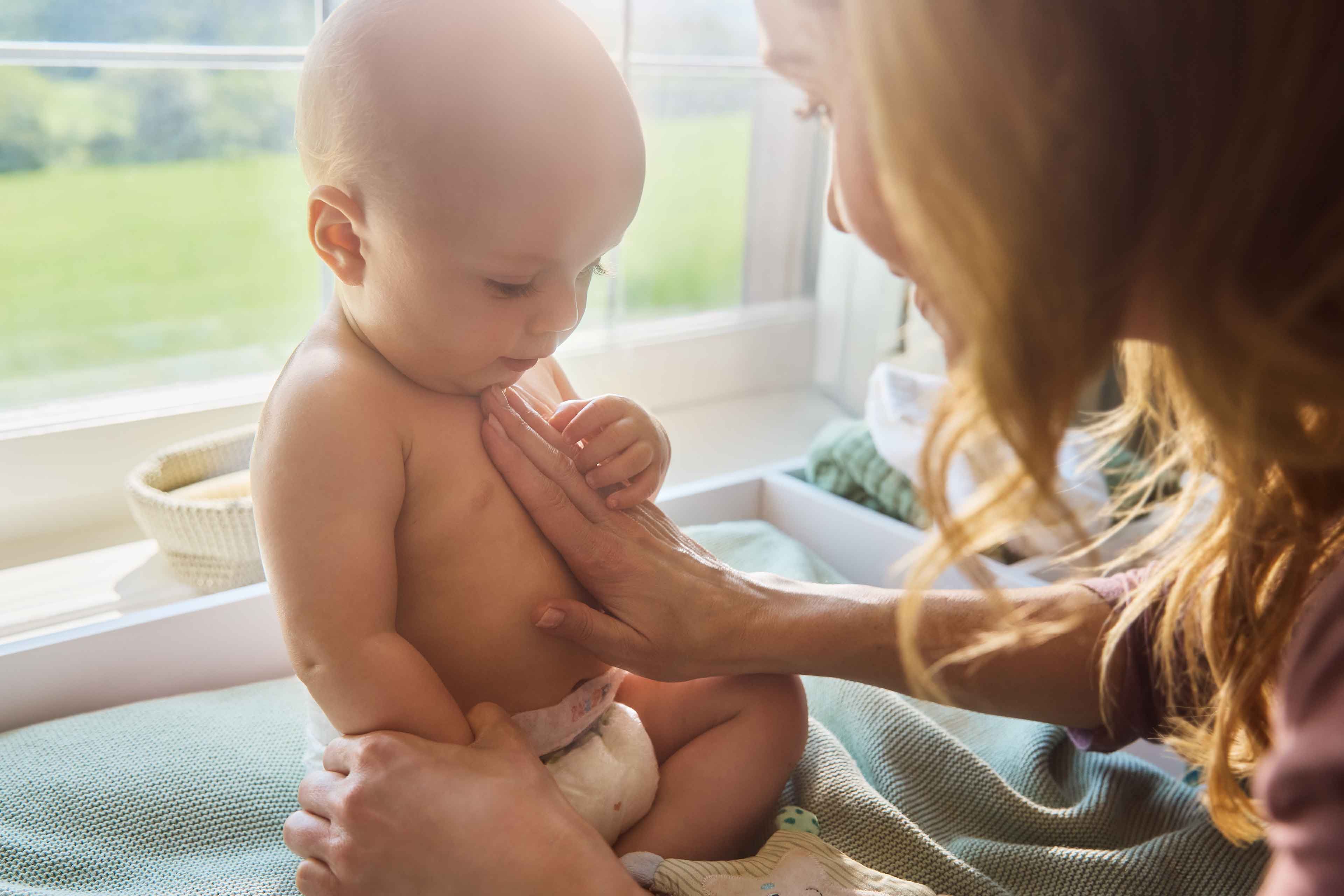 Mutter streichelt liebevoll die nackte, windeltragende Babybrust am Wickelplatz im warmen Sonnenlicht – sanfte Pflege für sensible Haut.